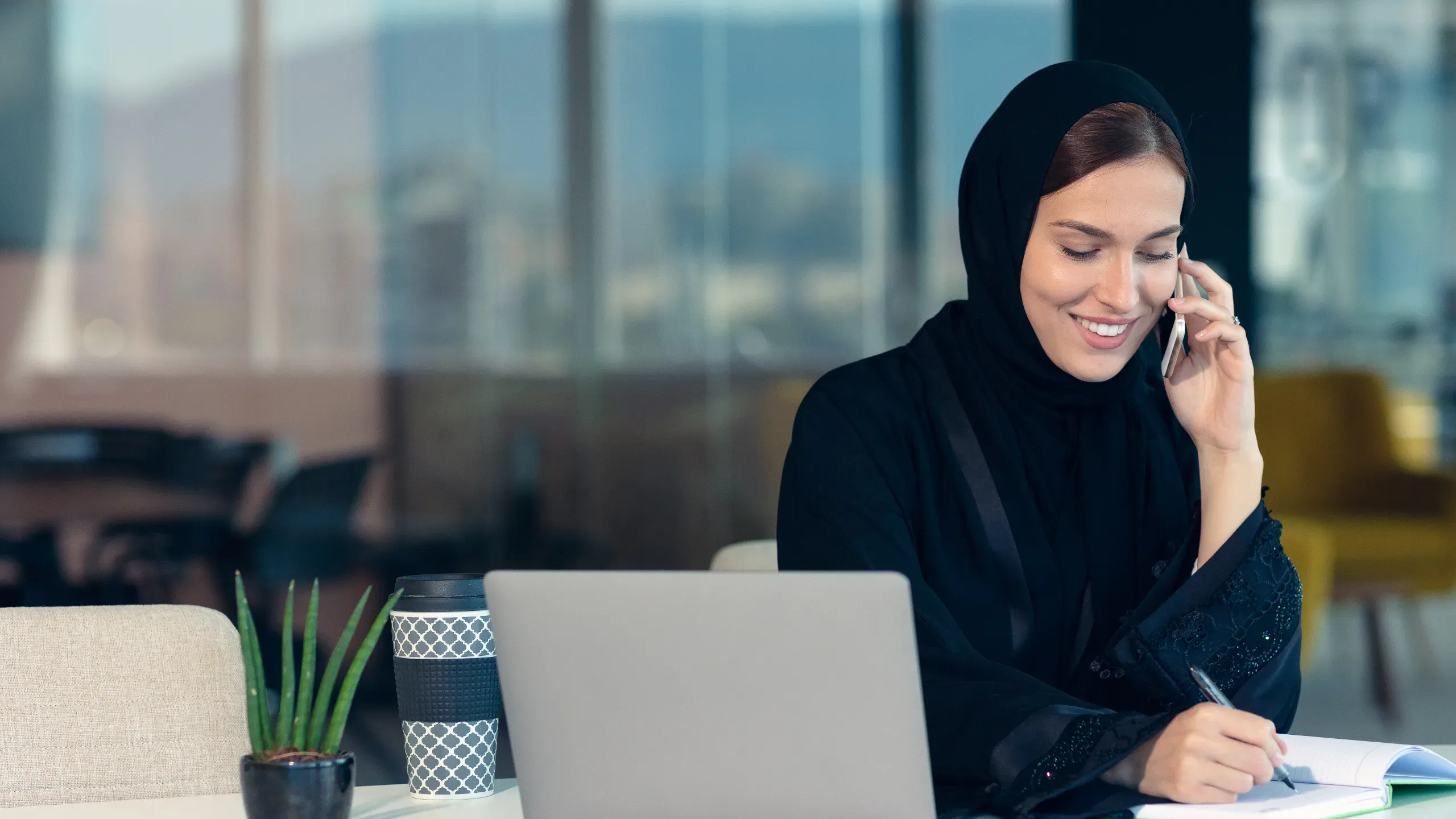 Middle Eastern businesswoman in abaya and hijab smiling, working on a laptop and phone in a modern office setting.